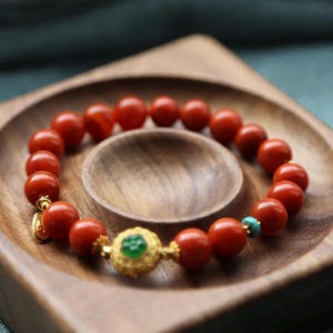 Puede incluir: Pulsera de cuentas rojas con un elemento central dorado adornado con una piedra verde y detalles turquesa. La pulsera se muestra sobre una bandeja de madera. Las cuentas miden aproximadamente 1 cm de di&aacute;metro.