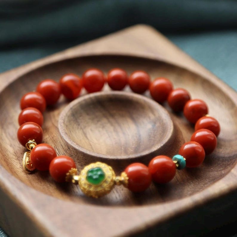 Puede incluir: Pulsera de cuentas rojas con un elemento central dorado adornado con una piedra verde y detalles turquesa. La pulsera se muestra sobre una bandeja de madera. Las cuentas miden aproximadamente 1 cm de di&aacute;metro.