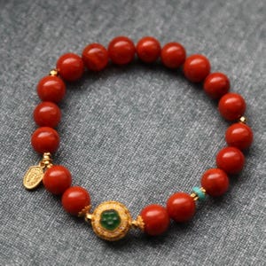 Puede incluir: Una pulsera de cuentas rojas con detalles dorados y un elemento decorativo central. La pulsera presenta cuentas redondas rojas pulidas, un dije dorado y una pieza central dorada con una piedra verde. Una peque&ntilde;a cuenta turquesa a&ntilde;ade un toque de color.