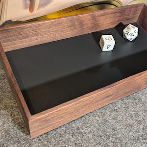 May include: A wooden dice tray with a dark stain and a black interior. Two white dice with black numbers are inside the tray. The tray is rectangular with angled sides, designed for tabletop gaming.