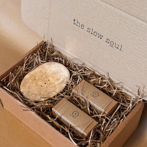Op de afbeelding: Een bruine kartonnen geschenkdoos met een rond, lichtgekleurd houten zeepbakje en twee rechthoekige, in bruin papier verpakte blokken. De binnenkant van het deksel bevat de tekst "the slow soul." De items liggen in een bed van snipperpapier.