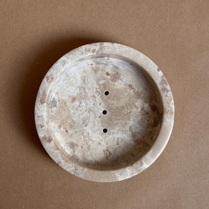 Könnte beinhalten: Eine runde, beige Seifenschale mit drei Abflusslöchern. Die Schale hat ein gesprenkeltes Muster mit hellen und dunklen Brauntönen. Der Rand ist leicht erhöht, und das Design ist schlicht und funktional.