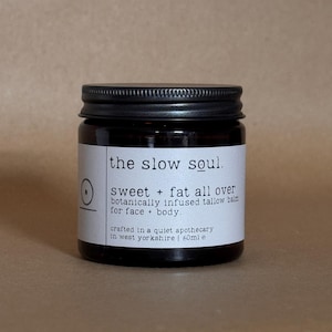 Op de afbeelding: Een donker amberkleurige glazen pot met een zwarte schroefdop en een wit etiket. Op het etiket staat: the slow soul. sweet + fat all over botanically infused tallow balm for face + body. crafted in a quiet apothecary in west yorkshire | 60ml e.