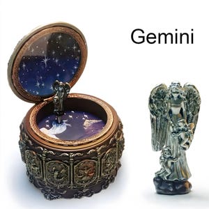 May include: A round, dark brown music box with a hinged lid featuring the text Gemini. The exterior displays relief carvings of zodiac signs. The interior lid and base show a blue starry night sky. A silver-toned angel figurine stands separately.