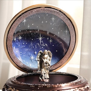 May include: A round, dark brown music box featuring a metallic angel figurine standing in the center. The lid is open, revealing a circular backdrop painted with a deep blue night sky, white stars, and a decorative bronze-toned frame border.
