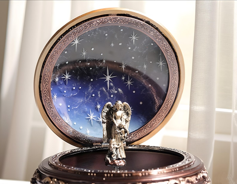 May include: A round, dark brown music box featuring a metallic angel figurine standing in the center. The lid is open, revealing a circular backdrop painted with a deep blue night sky, white stars, and a decorative bronze-toned frame border.