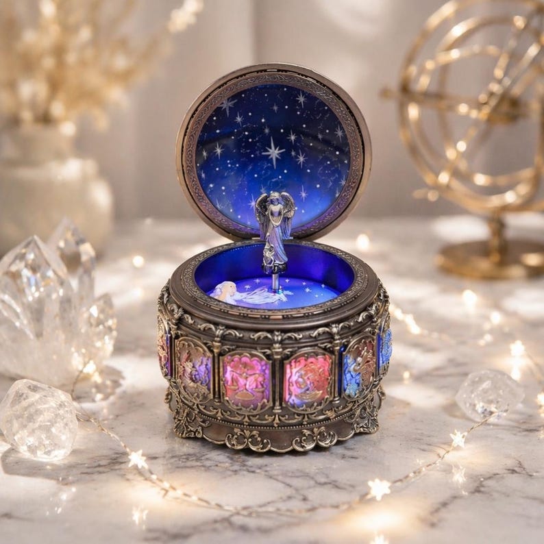May include: A round, metallic music box featuring an open lid with a dark blue starry sky interior. A silver angel figurine stands in the center. The exterior base is decorated with illuminated, colorful panels depicting zodiac signs.