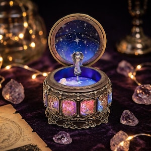 May include: A round, bronze-toned music box featuring a central silver-colored angel figurine. The lid interior displays a dark blue starry night sky. The exterior base is decorated with illuminated, colorful panels depicting zodiac signs.
