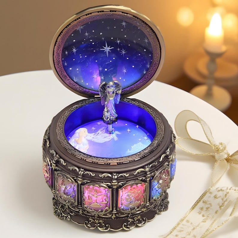May include: A round, dark-toned music box with an open lid revealing a central angel figurine. The interior features a blue-lit starry sky design. The exterior base is decorated with illuminated panels depicting zodiac symbols in various colors.