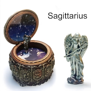 May include: A round brown music box with an open lid featuring a dark blue starry sky interior and a metallic angel figurine. The exterior displays zodiac relief carvings. Beside it stands a separate silver-toned angel figurine holding a bow.