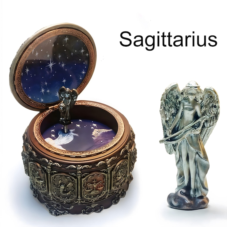 May include: A round brown music box with an open lid featuring a dark blue starry sky interior and a metallic angel figurine. The exterior displays zodiac relief carvings. Beside it stands a separate silver-toned angel figurine holding a bow.
