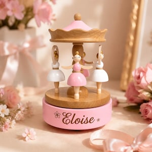 Può includere: Un carosello musicale in legno con tre statuette di ballerine in abiti bianchi e rosa. La base è dipinta di rosa chiaro e riporta il nome Eloise in corsivo, affiancato da due piccole icone di fiori su entrambi i lati.