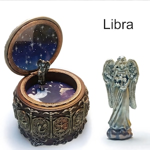 May include: A round brown music box with a hinged lid featuring a dark blue starry sky interior and zodiac relief panels on the exterior. A separate silver-toned angel figurine stands beside the box. The text Libra is printed in black.
