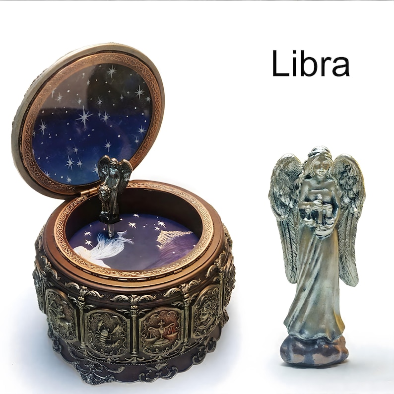 May include: A round brown music box with a hinged lid featuring a dark blue starry sky interior and zodiac relief panels on the exterior. A separate silver-toned angel figurine stands beside the box. The text Libra is printed in black.