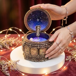 May include: A round, bronze-toned music box with an open lid revealing a central angel figurine. The interior features a dark blue starry sky design. The exterior of the box is decorated with embossed panels depicting zodiac signs in a metallic finish.