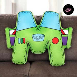 A plush toy in the shape of the letter "M" is sitting on a couch.
