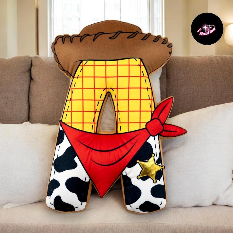 A plush toy of the letter "A" in the shape of a cowboy hat and pants, with a red bandana and a gold star, is sitting on a couch.
