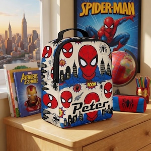 A Spider-Man themed lunch bag with the character Peter on it, placed on a wooden surface next to a Spider-Man poster and some books.