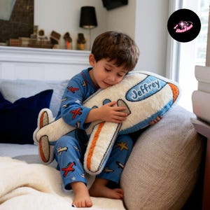A young boy is sitting on a couch, holding a stuffed airplane pillow with the name "Jeffrey" written on it.