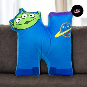 A plush pillow with a green alien character and a space-themed design, sitting on a couch.