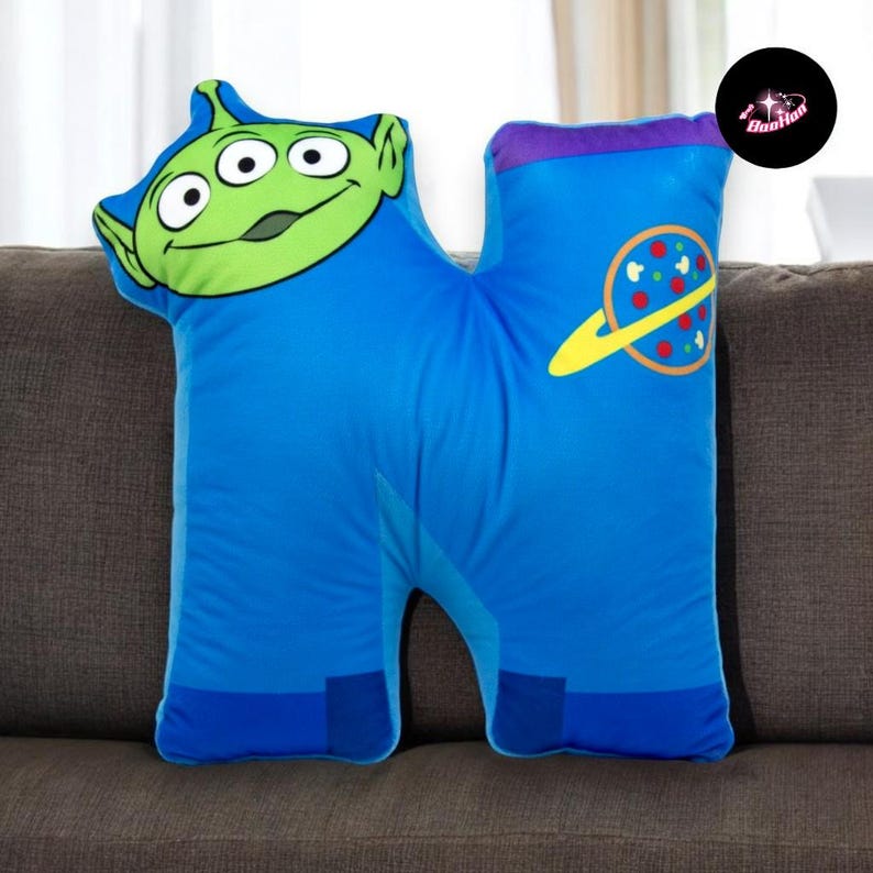 A plush pillow with a green alien character and a space-themed design, sitting on a couch.