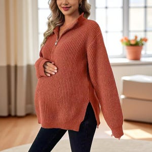 May include: Rust-colored maternity sweater with a ribbed texture and a half-zip closure. The sweater has long sleeves and side slits. The model is wearing black pants. The sweater is designed for comfort and style during pregnancy.