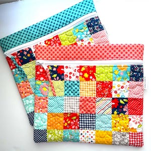 May include: Two square zippered pouches made from quilted fabric featuring a patchwork design of various colorful floral, geometric, and houndstooth patterns. Each pouch has a white zipper closure and a small white fabric tag labeled Quilted Comfort SC.