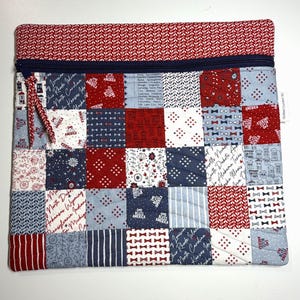 K&ouml;nnte beinhalten: Eine rechteckige, gesteppte Rei&szlig;verschlusstasche mit einem Patchwork-Design aus roten, wei&szlig;en und blauen Stoffquadraten. Die Muster zeigen Staatsnamen, kleine Fliegen, Sterne und Blumenmotive. Ein dunkelblauer Rei&szlig;verschluss verl&auml;uft oben.