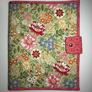 May include: A rectangular quilted fabric cover featuring a light green background with a dense floral pattern of pink, red, orange, and blue flowers and green leaves. The cover has a pink geometric border and a matching pink tab with a gold snap.