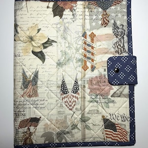 May include: Quilted fabric notebook cover with a patriotic theme. The cover features a collage of American flags, floral designs, and historical text. A navy blue border with white stars surrounds the cover, and a navy blue flap with a snap closure is on the right side.
