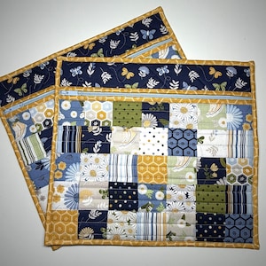May include: Two square quilted pouches featuring a patchwork design of blue, yellow, green, and white fabrics with floral, honeycomb, and polka dot patterns. Each pouch has a light blue zipper closure and a mustard yellow fabric border trim.