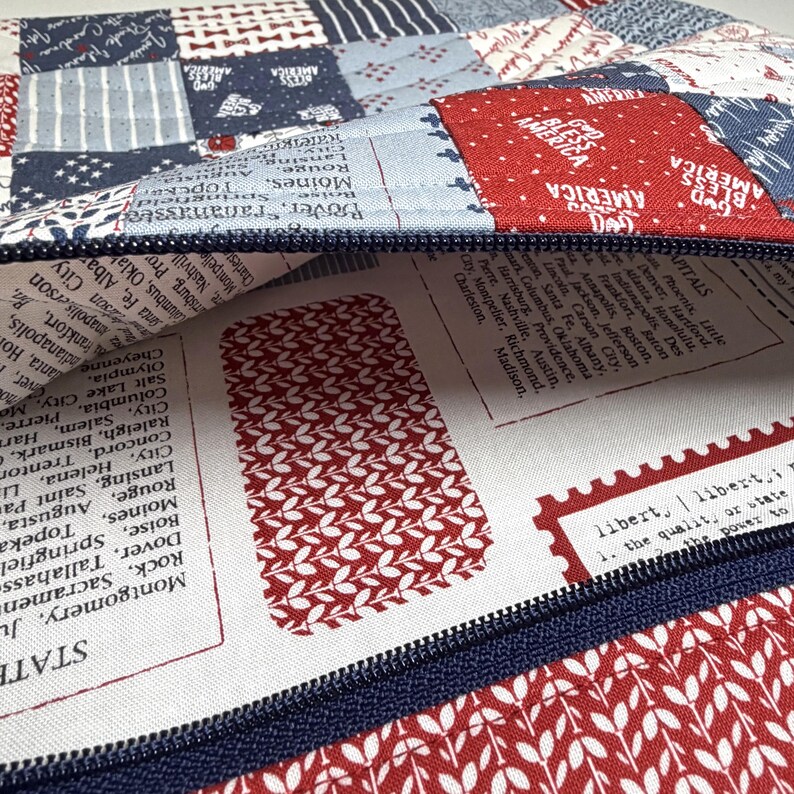 K&ouml;nnte beinhalten: Eine Rei&szlig;verschlusstasche mit einem Patchwork-Au&szlig;enmaterial aus roten, wei&szlig;en und blauen Stoffquadraten, die mit "God Bless America" und verschiedenen Mustern bedruckt sind. Das Innenfutter ist wei&szlig;er Stoff mit Listen von US-Hauptst&auml;dten und Text.