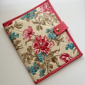 May include: Quilted fabric case with a floral pattern featuring large pink and red flowers, blue accents, and brown leaves on a cream background. The case has a pink border and a pink dotted flap with a gold snap closure.