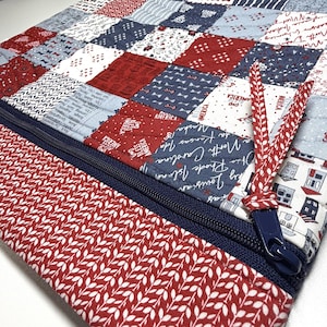 K&ouml;nnte beinhalten: Eine gesteppte Stofftasche mit einem Patchwork-Design in Rot, Wei&szlig; und Marineblau. Das Muster enth&auml;lt Texte wie "God Bless America" und verschiedene Staatsnamen. Ein dunkelblauer Rei&szlig;verschluss mit einem rot-wei&szlig; gemusterten Stoffanh&auml;nger.