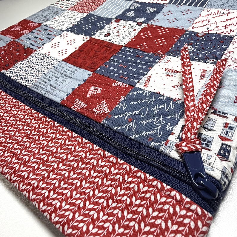K&ouml;nnte beinhalten: Eine gesteppte Stofftasche mit einem Patchwork-Design in Rot, Wei&szlig; und Marineblau. Das Muster enth&auml;lt Texte wie "God Bless America" und verschiedene Staatsnamen. Ein dunkelblauer Rei&szlig;verschluss mit einem rot-wei&szlig; gemusterten Stoffanh&auml;nger.