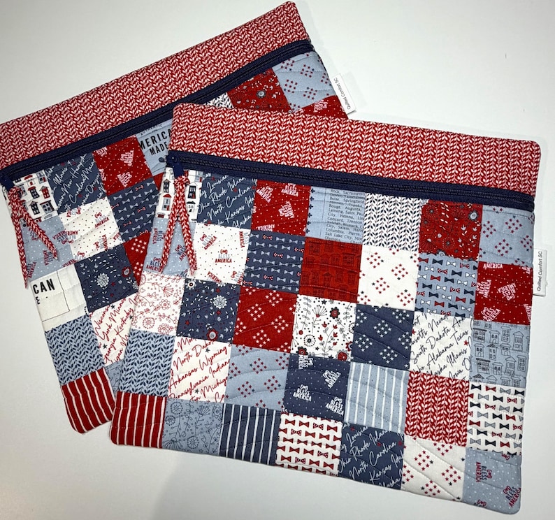 K&ouml;nnte beinhalten: Zwei rechteckige, gesteppte Rei&szlig;verschlusstaschen mit einem Patchwork-Design in Rot, Wei&szlig; und Blau. Die Stoffmuster zeigen Staatsnamen, kleine Fliegen, Blumenmotive und den Schriftzug America. Jede Tasche hat einen dunkelblauen Rei&szlig;verschluss.