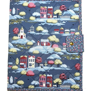 May include: A quilted fabric book cover featuring a blue background with a repeating pattern of houses, trees, and small figures. The cover has a floral-patterned border and a matching tab closure secured with a single gold-toned metal snap.