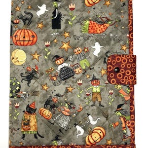 May include: A quilted fabric cover featuring a gray background with a Halloween-themed print of witches, pumpkins, ghosts, black cats, and stars. The cover has a dark orange border with a repeating circle pattern and a matching snap-button closure.