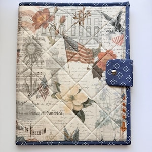 Può includere: Una copertina in tessuto trapuntato con un collage di immagini patriottiche tra cui la Casa Bianca, bandiere americane, fiori di magnolia e testo in corsivo. La copertina ha un bordo blu scuro con stelle bianche e una linguetta di chiusura.