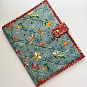 May include: Quilted fabric notebook cover with a floral pattern in shades of blue, yellow, and red. The cover features a button closure and a contrasting red floral border. The design includes flowers, ladybugs, and butterflies.