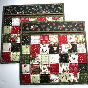 May include: Two Christmas-themed quilted placemats. Each placemat features a patchwork design with red, green, and white squares. The top border has a black fabric with white snowflakes and red floral accents. The edges are trimmed with a green fabric.