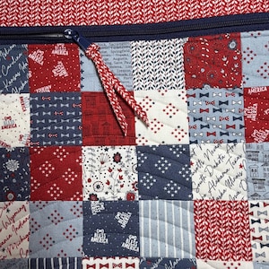 K&ouml;nnte beinhalten: Eine gesteppte Stofftasche mit einem Patchwork-Design in Rot, Wei&szlig; und Blau. Der Stoff zeigt Muster mit US-Bundesstaatsnamen, dem Text "God Bless America", kleinen Fliegen und Blumenmotiven. Ein dunkelblauer Rei&szlig;verschluss mit Kordelzug ist zu sehen.
