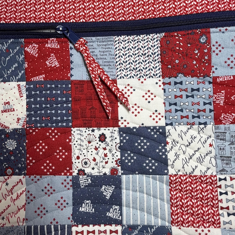 K&ouml;nnte beinhalten: Eine gesteppte Stofftasche mit einem Patchwork-Design in Rot, Wei&szlig; und Blau. Der Stoff zeigt Muster mit US-Bundesstaatsnamen, dem Text "God Bless America", kleinen Fliegen und Blumenmotiven. Ein dunkelblauer Rei&szlig;verschluss mit Kordelzug ist zu sehen.