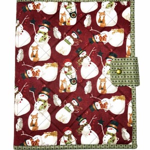 May include: A quilted fabric book cover featuring a dark red background with a repeating pattern of snowmen, foxes, and hedgehogs. The cover includes a green fabric trim with a star pattern and a matching side tab secured with a brass-colored snap.