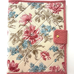 Könnte beinhalten: Gesteppte Notizbuchhülle mit floralem Muster. Der cremefarbene Hintergrund zeigt große rosa und blaue Blumenmuster. Mit einem rosa Rand und einem passenden rosa Verschluss mit goldenem Knopf. Die Notizbuchhülle ist rautenförmig gesteppt.