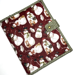 May include: A quilted fabric cover featuring a dark red background with a repeating pattern of snowmen, foxes, and hedgehogs. The edges are trimmed with a green fabric featuring a small star pattern, and a matching tab with a gold snap closure.