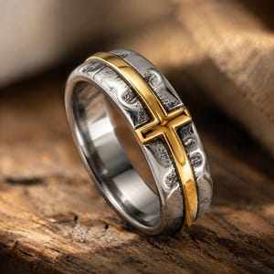 May include: A two-tone ring featuring a textured, dark silver-coloured band with a polished gold-coloured cross inlay that wraps around the centre. The band has a rugged, hammered finish on the outer surface and a smooth, reflective interior.