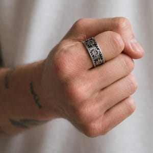 May include: A wide silver-toned metal ring featuring a repeating pattern of raised gear or cogwheel motifs around the entire band. The surface of the ring has a textured, darkened finish within the recessed areas to highlight the mechanical design.