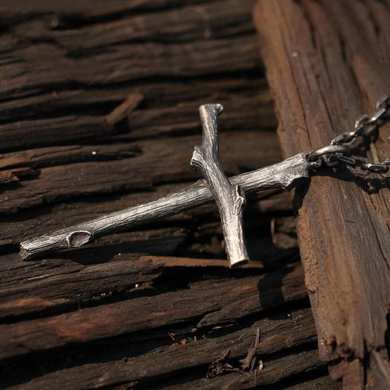 May include: A silver-colored pendant necklace featuring a cross design crafted to resemble intertwined branches. The cross is detailed with textured surfaces and is suspended from a delicate chain. The necklace rests on a weathered wooden surface.