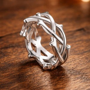May include: A silver-colored ring with a unique design. The ring features an interwoven pattern of bone-like structures, creating an intricate and eye-catching aesthetic. The ring is displayed on a wooden surface.